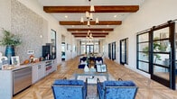 Located at 15901 Championship Pkwy in Fort Worth, TX, this single-story clubhouse offers a generously sized, open-plan living space designed with a modern farmhouse aesthetic. The interior is immediately striking, with soaring ceilings adorned by rich, exposed wooden beams that run the length of the room, complemented by elegant pendant chandeliers that provide warm, layered lighting throughout. The floors feature a distinctive geometric parquet wood pattern, adding visual depth and sophistication to the expansive layout. Crisp white shiplap walls serve as a bright, clean backdrop, enhancing the sense of openness and allowing curated artwork and sculptural wall pieces to stand out prominently. The living area is thoughtfully arranged into multiple functional zones, including comfortable lounge seating with plush blue velvet and neutral upholstered sofas, a dining area with wood tables and upholstered chairs, and a beverage station along one wall. Black-framed glass French doors and large windows flood the interior with natural light while offering views of the outdoor surroundings. A sliding barn-style glass door adds an architectural detail that bridges indoor spaces seamlessly. The overall ambiance is refined yet inviting, balancing contemporary design with warm, rustic touches.