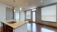 This well-appointed single-floor space showcases a thoughtful modern design with high-quality finishes throughout. The open-concept living and kitchen area is anchored by rich, dark hardwood flooring that flows seamlessly across the main living spaces. The kitchen is a standout feature, equipped with full-height dark espresso cabinetry, a full suite of stainless steel appliances including a refrigerator, range, and built-in microwave, and a large granite-topped island with an undermount sink and pendant lighting above. Recessed lighting throughout the main area ensures a well-lit environment at all times. The living area benefits from multiple floor-to-ceiling windows fitted with horizontal blinds, allowing for ample natural light and a bright, airy atmosphere. A ceiling fan adds comfort to the space. The bedroom offers plush carpeting, a generous window with outdoor views, and a clean, neutral palette. The bathroom is finished with large-format tile flooring, dark wood vanity cabinetry, a granite countertop with a modern faucet, a framed mirror, and a tiled tub-shower combination. The overall ambiance is contemporary and refined, combining practical functionality with stylish, durable materials.