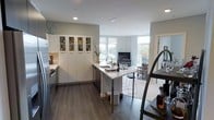 This single-floor space showcases a thoughtfully designed open-concept layout that integrates the kitchen and living areas into one cohesive environment. The kitchen is a standout feature, anchored by a central island with a light-toned countertop that doubles as a casual dining and entertaining surface, complete with bar-height seating. Crisp white cabinetry lines the walls, including a tall display cabinet with glass-front upper sections, ideal for showcasing glassware and bottles. Stainless steel appliances add a contemporary, functional touch, while dark hardwood-style flooring grounds the space with warmth and contrast against the white palette. Recessed lighting and a pendant fixture above the island provide layered illumination throughout. The kitchen transitions naturally into the living area, where large windows on multiple walls invite abundant natural light, creating an airy, open atmosphere. The living space is oriented around a media unit and features a light-toned area rug that defines the seating zone. The overall design balances clean, modern aesthetics with practical functionality, making it a well-appointed and versatile space.