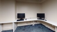 This single-floor business center is thoughtfully designed to support a range of professional and computing needs. The space is anchored by a large, L-shaped white built-in desk that wraps around the corner of the main work area, providing ample surface space for multiple users simultaneously. Two desktop computer workstations with monitors and keyboards are positioned along the desk, making the area well-suited for tasks requiring focused computer use. An additional dedicated workstation with its own monitor and keyboard occupies a separate alcove, offering a more private computing environment. A wall-mounted Samsung flat-screen television is installed above one section of the desk, accompanied by a media device and remote, adding a multimedia element to the space. An overhead wall-mounted cabinet provides supplementary storage, while a two-drawer lateral filing cabinet in a rich wood finish adds both function and a refined aesthetic. Artwork featuring soft, abstract brushstrokes in muted tones is framed and displayed on the wall, contributing warmth to the otherwise neutral palette of cream-toned walls and white trim. Blue patterned carpet tiles cover the floor throughout, adding texture and visual interest underfoot. A glimpse into an adjoining lounge area reveals tiled flooring, a blue leather accent chair, and warm ambient lighting, suggesting a comfortable space for relaxation nearby.