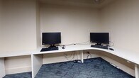 This single-floor business center is thoughtfully designed to support a range of professional and computing needs. The space is anchored by a large, L-shaped white built-in desk that wraps around the corner of the main work area, providing ample surface space for multiple users simultaneously. Two desktop computer workstations with monitors and keyboards are positioned along the desk, making the area well-suited for tasks requiring focused computer use. An additional dedicated workstation with its own monitor and keyboard occupies a separate alcove, offering a more private computing environment. A wall-mounted Samsung flat-screen television is installed above one section of the desk, accompanied by a media device and remote, adding a multimedia element to the space. An overhead wall-mounted cabinet provides supplementary storage, while a two-drawer lateral filing cabinet in a rich wood finish adds both function and a refined aesthetic. Artwork featuring soft, abstract brushstrokes in muted tones is framed and displayed on the wall, contributing warmth to the otherwise neutral palette of cream-toned walls and white trim. Blue patterned carpet tiles cover the floor throughout, adding texture and visual interest underfoot. A glimpse into an adjoining lounge area reveals tiled flooring, a blue leather accent chair, and warm ambient lighting, suggesting a comfortable space for relaxation nearby.