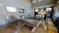 This sophisticated entertainment lounge is thoughtfully designed to offer a premium social experience across a single, open floor. The space immediately impresses with its soaring double-height ceilings and beautifully paneled walls finished in a warm neutral tone, lending a refined yet welcoming atmosphere throughout.

At the heart of the room, two custom billiard tables with rich wood frames and grey felt tops take center stage atop gleaming hardwood floors, making them natural focal points for recreation and social gathering. Multiple wall-mounted flat-screen televisions are strategically positioned around the space, ensuring excellent viewing angles from virtually every vantage point.

Toward the rear, a stylish bar area anchors the room with pendant lighting, a decorative tile backsplash, and a row of sleek white upholstered bar stools. The bar is equipped with shelving and display screens above, creating an inviting hub for refreshments and conversation.

Complementing the bar, a seating area with high-top tables and bar chairs offers additional space for guests to relax. The overall ambiance strikes a balance between upscale lounge and lively game room, with warm lighting, cohesive finishes, and a well-considered layout that encourages both active play and casual socializing.