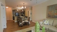 This single-story home showcases an open-concept layout that seamlessly connects the kitchen, dining, and living areas into one cohesive and functional space. The kitchen is outfitted with dark cabinetry, a kitchen island with bar seating, and black appliances including a refrigerator, microwave, and range. A track lighting system illuminates the workspace efficiently. Adjacent to the kitchen, the dining area is centered beneath an elegant chandelier with warm-toned Edison-style bulbs, creating an inviting atmosphere for gatherings. The space comfortably accommodates a round glass dining table surrounded by woven rattan chairs with white tie accents. Flowing naturally from the dining area, the living space features neutral beige walls, warm hardwood flooring throughout, and a plush area rug that anchors the seating arrangement. Crown molding runs along the ceiling, adding a refined architectural detail. Decorative green lantern accents and coordinating throw pillows introduce pops of color. The warm, neutral palette combined with consistent flooring and thoughtful lighting gives the entire space a cohesive, welcoming character.