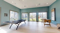 This well-appointed game room in Terrell, TX, is designed to foster recreation and community engagement in a welcoming, modern setting. The space is finished with large-format light tile flooring that reflects abundant natural light streaming in through multiple oversized sliding glass doors and wide windows. These openings frame views of the surrounding outdoor grounds and neighboring buildings, creating a bright and connected atmosphere throughout the day.

The teal-painted walls lend a fresh, energetic character to the room, complemented by recessed ceiling lighting that ensures consistent illumination. A ping-pong table anchors the center of the space, while two rustic wood foosball tables flank the entrance, adding a warm, textural contrast to the contemporary surroundings. Framed abstract artwork decorates the walls near the entry double doors, contributing an artistic touch to the recreational environment.

Along one wall, a built-in bar-height counter with seating and warm wall sconces creates a comfortable lounge zone, accompanied by a wall-mounted flat-screen television — ideal for spectators or casual gatherings. The space connects through glass-paneled double doors to adjacent common areas. The overall design balances function and style, making this game room a well-equipped and inviting hub for leisure and social activity.
