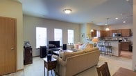This two-story home offers a thoughtfully designed layout that prioritizes open, connected living. The main level centers around a spacious open-concept living area, where the living room, dining space, and kitchen flow naturally into one another. The living room is comfortably sized, featuring plush carpeted flooring, multiple windows that invite an abundance of natural light, and ample space for seating and entertainment. A large sectional sofa anchors the room, complemented by a media setup and surrounding audio equipment. Adjacent to the living area, the kitchen is well-equipped with warm wood cabinetry, pendant lighting, and a raised breakfast bar with barstool seating — ideal for casual dining or entertaining. The open sightlines between the kitchen and living room create a welcoming, social atmosphere throughout the main level. Neutral tones and soft lighting contribute to a warm and inviting ambiance throughout. The second floor provides additional living space, offering privacy and versatility. Overall, this home presents a practical and comfortable layout suited for everyday living, with a design that balances openness on the main level with defined spaces across both floors.