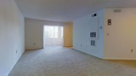 This single-floor residential space offers a clean, functional layout designed for comfortable everyday living. The living area serves as the heart of the home, featuring soft wall-to-wall carpeting that adds warmth underfoot and a neutral color palette throughout that creates a calm, inviting atmosphere. A full-height sliding glass door, dressed with vertical blinds, fills the room with abundant natural light while providing direct access to an outdoor space — ideal for enjoying fresh air and exterior views. The white walls and smooth ceilings keep the interior feeling open and airy. The space is equipped with built-in HVAC vents and a wall-mounted thermostat, ensuring efficient climate control in all seasons. Additional wall vents provide consistent airflow distribution throughout the living area. The understated design aesthetic, combined with practical amenities, makes this a versatile space well-suited to a variety of furnishing arrangements and personal styles. The overall ambiance is bright, neutral, and move-in ready, offering a solid foundation for personalizing into a comfortable home.