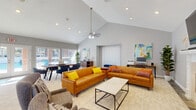 This single-floor clubhouse is designed to serve as a versatile community gathering space, highlighted by dramatic vaulted ceilings that lend an airy, expansive feel throughout. The main lounge area features two warm caramel leather sofas accented with vibrant throw pillows, centered around a marble-top coffee table and anchored by a tile-surround fireplace with a wall-mounted television above. A ceiling fan circulates air through the generous open layout, while recessed lighting and colorful abstract artwork add a polished, contemporary touch.

Adjacent to the lounge, a dedicated dining area accommodates a large wooden table with navy upholstered chairs, positioned near floor-to-ceiling windows and French doors that open directly to an outdoor pool area, flooding the space with abundant natural light. A workstation area with a computer and printer provides a functional resource corner for residents.

The kitchen features dark wood cabinetry, stone-tile backsplash, black appliances, and a granite-topped island with seating for several teal velvet bar stools. Stone tile flooring anchors the kitchen zone, transitioning into carpeted areas throughout the rest of the space. Together, these areas create a well-rounded, inviting environment suitable for socializing, working, and entertaining.