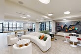 Located at 3100 Lake Villa Dr in Metairie, Louisiana, this single-floor amenity space offers a well-appointed community lounge and recreation area designed with a clean, contemporary aesthetic. The open-concept layout is anchored by light herringbone-patterned tile flooring and illuminated throughout by distinctive circular flush-mount ceiling fixtures that create a bright, welcoming atmosphere.

The main lounge area features a curved white sofa accented with green and orange throw pillows, paired with swivel accent chairs and terrazzo and wood side tables. A bold, hexagonal decorative shelving unit mounted on a wood-paneled accent wall serves as a striking focal point, displaying colorful books, plants, and decorative objects. A dark marble-top dining table with modern gold-base stools provides additional seating nearby.

The game room zone is equipped with a pool table and a ping-pong table, with large floor-to-ceiling windows that flood the space with natural light and offer views of the surrounding building exterior. Wall-mounted flat-screen televisions are positioned throughout for entertainment.

Adjacent to the lounge, a fully equipped fitness center is enclosed behind sleek floor-to-ceiling glass walls and black-framed doors, housing cardio machines and free weights. The transparent enclosure maintains the open, airy feel of the overall space while clearly delineating the workout area.