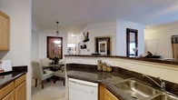 This single-floor residence showcases a thoughtfully designed open-concept layout that seamlessly connects the kitchen and dining area. The kitchen is outfitted with rich dark granite countertops that wrap around the workspace, providing ample surface area for meal preparation. Warm wood cabinetry complements the stone surfaces, while a stainless steel sink and white dishwasher add functional, classic touches. A low breakfast bar-style counter serves as a natural divider between the kitchen and the adjacent dining space, maintaining an open and airy feel throughout.

Crown molding runs along the ceiling, adding an elegant architectural detail that elevates the overall aesthetic. The dining area accommodates a round dark wood table surrounded by cushioned chairs, lit by a decorative pendant light fixture that creates a warm, intimate atmosphere. Framed artwork with ornate dark frames adorns the walls, contributing to a refined, traditional style. Light-toned flooring and white walls reflect natural and artificial light effectively, enhancing the sense of space. The overall ambiance is warm, polished, and well-coordinated, with consistent design elements tying each area together cohesively.