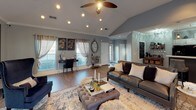 This single-story clubhouse offers a thoughtfully designed open-concept layout that seamlessly connects its key gathering areas. The living room serves as the centerpiece, featuring vaulted ceilings with recessed lighting, a ceiling fan, and crown molding. A white-mantled fireplace with a mosaic tile surround anchors one wall, complemented by wood-look flooring that flows throughout the space. Large windows dressed with patterned sheer curtains allow generous natural light to fill the room, creating a bright and welcoming atmosphere.

The kitchen is well-appointed with white cabinetry, light countertops, and a gray geometric tile backsplash. Black appliances, including a side-by-side refrigerator with an ice and water dispenser, an electric range, and a microwave, provide a bold contrast to the lighter cabinetry. A breakfast bar with stylish upholstered counter-height seating connects the kitchen to the main gathering area, making it ideal for community events and social gatherings.

A separate dining area accommodates a large wooden table surrounded by navy upholstered chairs, with a window providing an outdoor view. The overall ambiance is sophisticated and inviting, with consistent cool-toned gray walls tying each area together cohesively.