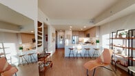 This single-floor residence showcases a thoughtfully designed open-concept layout that seamlessly connects the living, dining, and kitchen areas. The kitchen is a standout feature, outfitted with light wood cabinetry extending to the ceiling, stainless steel appliances including a gas range and built-in microwave, and a generous white quartz island with bar seating for four. Recessed lighting and under-cabinet illumination keep the space bright and functional throughout.

The living area benefits from abundant natural light streaming through expansive floor-to-ceiling windows, which frame sweeping city skyline views. The space accommodates a relaxed, curated layout with room for lounge seating and open shelving.

The bedroom is a particular highlight, featuring a dramatic corner window installation that offers panoramic urban views and fills the room with warm natural light. The bathroom is finished with a floating double-sink vanity in light wood tones, a large backlit mirror, white quartz countertops, and a glass-enclosed shower with neutral tile surround.

Throughout the residence, warm wood-look flooring ties each area together, complementing the clean white walls and creating a cohesive, contemporary aesthetic with a welcoming, residential warmth.