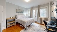 Located at 50-52 Irving St in Cambridge, MA, this one-bedroom, one-bathroom unit offers a well-organized and efficiently designed living space across a single floor. The bedroom is a bright, inviting room featuring warm hardwood floors, soft neutral-toned walls, and abundant natural light streaming through multiple windows and French doors that open to an outdoor area. A classic cast-iron radiator adds a charming, period-appropriate detail, while crown molding lends a refined architectural touch. The room accommodates a workspace area with open shelving, a clothing rack, and wardrobe storage, making it highly functional. The open living area flows naturally from the bedroom, incorporating a dedicated desk zone and access to a compact kitchen with essentials visible in the background. Exposed copper pipe accents along the ceiling add an industrial character to the otherwise clean, neutral aesthetic. The bathroom features a sleek, glass-enclosed walk-in shower finished with crisp white subway tiles, hexagonal mosaic floor tiles, and brushed nickel fixtures, delivering a modern and polished look. Throughout the unit, hardwood flooring, crown molding, and a cohesive light gray color palette create a consistent, comfortable ambiance suited for everyday living in the heart of Cambridge.