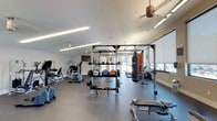 This single-floor fitness center is designed to support a comprehensive range of workout routines in a clean, modern environment. The space is organized into distinct training zones, each thoughtfully outfitted to meet diverse fitness needs. The strength training area features a full power rack, multiple weight machines, adjustable benches, barbells, and a well-stocked rack of medicine balls and weight plates, providing everything needed for resistance-based workouts. A dedicated functional training zone includes an Everlast heavy punching bag, battle ropes, and cable attachments, ideal for high-intensity and combat-style training. The cardio section lines the perimeter near large windows fitted with roller shades, housing treadmills, ellipticals, a stationary bike, and a rowing machine — all bathed in natural light that creates an energizing atmosphere. Throughout the space, durable rubber flooring provides safety and comfort underfoot, while recessed linear LED lighting and ceiling fans ensure a well-lit, comfortable environment. A wall-mounted television adds to the amenity-rich setting. White walls and high ceilings contribute to an open, airy feel, and direct outdoor access through glass doors enhances the sense of connectivity to the surrounding environment.