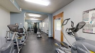 Located at Northwood Drive in Eagan, Minnesota, this fitness center offers a comprehensive workout facility designed to accommodate a range of exercise needs. The space is laid out in a long, elongated floor plan that flows efficiently from one zone to the next, connected by an arched passageway that divides the cardio and strength training areas.

The main cardio section features multiple treadmills and elliptical trainers arranged in a row, complemented by full-length wall mirrors that visually expand the space and allow users to monitor their form. Flush-mounted fluorescent lighting and a ceiling fan provide consistent illumination and airflow throughout. Windows fitted with horizontal blinds allow natural light to filter in while maintaining privacy.

Through the arched opening, the strength training area is equipped with a multi-station cable machine, a flat weight bench, and a dumbbell rack stocked with a range of free weights. Exercise charts are mounted on the walls for easy reference. Durable rubber flooring covers the entire facility, offering both safety and easy maintenance underfoot.

The space features a textured ceiling, neutral grey and blue-accented walls, and a private entry door with a windowed panel framed in warm wood trim. A coat rack near the entrance adds a practical touch. The overall ambiance is clean, functional, and well-organized, making it a practical fitness resource for the surrounding community.