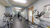Located at Northwood Drive in Eagan, Minnesota, this fitness center offers a comprehensive workout facility designed to accommodate a range of exercise needs. The space is laid out in a long, elongated floor plan that flows efficiently from one zone to the next, connected by an arched passageway that divides the cardio and strength training areas.

The main cardio section features multiple treadmills and elliptical trainers arranged in a row, complemented by full-length wall mirrors that visually expand the space and allow users to monitor their form. Flush-mounted fluorescent lighting and a ceiling fan provide consistent illumination and airflow throughout. Windows fitted with horizontal blinds allow natural light to filter in while maintaining privacy.

Through the arched opening, the strength training area is equipped with a multi-station cable machine, a flat weight bench, and a dumbbell rack stocked with a range of free weights. Exercise charts are mounted on the walls for easy reference. Durable rubber flooring covers the entire facility, offering both safety and easy maintenance underfoot.

The space features a textured ceiling, neutral grey and blue-accented walls, and a private entry door with a windowed panel framed in warm wood trim. A coat rack near the entrance adds a practical touch. The overall ambiance is clean, functional, and well-organized, making it a practical fitness resource for the surrounding community.