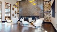 Located at 8555 Belle Dr in the Highlands Ranch community, this single-floor amenity space offers an impressive blend of modern design and multifunctional areas spanning a generous footprint. The main lounge greets visitors with soaring double-height ceilings, floor-to-ceiling windows that flood the space with natural light, and elegant cream-upholstered seating arranged around a gold-accented coffee table. Striking pendant lights with gold interiors add warmth and visual interest overhead. Adjacent to the lounge, a co-working and social area features rich dark hardwood-style flooring, exposed wood beam ceiling details, and a dramatic diamond-pattern bookshelf wall display. A large-scale wall mural map serves as a bold artistic focal point, complemented by long communal tables and bar-height seating ideal for work or casual gatherings. A dedicated indoor golf simulator room offers a full-turf hitting surface and a large projection screen, providing a premium recreational amenity. The kitchen area is appointed with a substantial island topped in white quartz, wood cabinetry, gold-accented bar stools, and large drum pendant lights, all framed by views of the outdoor pergola area through oversized windows. Throughout, the design maintains a cohesive contemporary aesthetic with curated décor and high-end finishes.