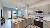 This single-floor residence showcases a thoughtfully designed open-concept layout that seamlessly connects the kitchen and living areas. The kitchen is a standout feature, equipped with stainless steel appliances including a range, microwave, refrigerator, and dishwasher. Gray shaker-style cabinetry pairs elegantly with granite countertops, while a classic white subway tile backsplash adds a clean, modern touch. A peninsula with seating provides additional workspace and casual dining, illuminated by globe pendant lights that add a contemporary flair. The living area offers a comfortable, light-filled space with neutral tones and natural light streaming through slatted blinds. The bedroom is a tranquil retreat featuring light wood-style flooring, a window with natural light, and a frosted-glass interior door for added privacy. An adjoining walk-in closet provides generous storage. The bathroom continues the modern aesthetic with white subway tile surrounding the tub and shower, a granite-topped vanity with gray cabinetry, and warm wood-style flooring. Throughout the residence, black matte hardware and fixtures provide consistent, contemporary detailing against the soft gray palette.