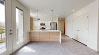 This thoughtfully designed single-floor unit presents a clean, contemporary aesthetic throughout. The heart of the home is an open-concept kitchen and living area bathed in natural light. The kitchen features a large central island with warm wood-toned cabinetry and a crisp white countertop, complemented by white upper cabinets, stainless steel appliances, and stylish pendant lighting. The open layout flows seamlessly into the living area, which offers direct access to a private balcony through a set of French doors, framing views of the surrounding neighborhood and distant hills.

Two carpeted bedrooms provide comfortable retreats, each featuring large windows that invite abundant natural light and neighborhood views. The neutral carpet and bright white walls create a calm, versatile backdrop.

The unit includes two bathrooms. The primary bathroom is equipped with a glass-enclosed walk-in shower, a single-sink vanity with wood-toned cabinetry, a large mirror, and neutral-toned flooring. A second bathroom features a clean, white soaking tub with subway tile surround. Throughout the unit, a consistent palette of soft whites, warm wood accents, and neutral flooring ties the spaces together into a cohesive, modern living environment.