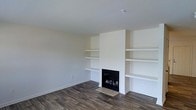 This two-floor space is designed with a clean, modern aesthetic that emphasizes simplicity and functionality. The main living area serves as a focal point, showcasing a built-in fireplace framed by symmetrical open shelving units on either side. These shelves extend from floor to ceiling on both flanks of the fireplace surround, offering generous display and storage space while creating a structured, architectural feel.

The living area features dark hardwood-style flooring that contrasts sharply with the bright white walls and ceiling, lending warmth and visual depth to the room. The neutral color palette throughout creates a calm, versatile backdrop suited to a variety of interior styles. The smooth, white walls are free of ornamentation, keeping the focus on the architectural built-ins and fireplace.

The fireplace itself is set into a broad, floor-to-ceiling white surround, adding a sense of height and grandeur to the space. A smoke detector is visible on the ceiling, indicating modern safety features are in place. Overall, the space presents a well-balanced, minimalist design that feels both welcoming and practical across its two levels.