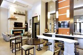 This thoughtfully designed single-floor amenity space blends contemporary style with inviting comfort. The main gathering area showcases light hardwood flooring, soaring ceilings, and an open layout that encourages both casual mingling and relaxed lounging. A sleek white curved bar counter lined with modern black stools anchors one side of the room, complemented by high-top bistro tables and ladder-back chairs — ideal for socializing or working.

A dramatic floor-to-ceiling black-framed glass garage door wall serves as a striking architectural feature, seamlessly connecting the interior to an adjacent outdoor courtyard. Double glass entry doors frame views of a charming brick-accented exterior with arched detailing, flooding the space with natural light.

The lounge area offers a warmer, more intimate atmosphere centered around a full-height stacked stone fireplace wall featuring an integrated linear electric fireplace with color-changing LED ambient lighting. A large flat-screen television is mounted above a rustic wood mantel, while built-in shelving adds functional display space. Plush seating, a patterned area rug, and curated artwork complete the refined yet relaxed ambiance, making this space as functional as it is visually compelling.