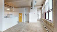 This single-floor space showcases a compelling blend of industrial and modern design elements. The concrete ceiling with exposed ductwork and ventilation systems lends an authentic industrial character, while the crisp white walls provide a clean, versatile backdrop throughout. The open main area is finished with neutral wall-to-wall carpeting, creating a softer contrast against the raw architectural details overhead.

A striking original brick wall runs along one side of the space, adding texture and a sense of history to the interior. Large windows along this same wall allow natural light to filter in, illuminating the space and highlighting its unique character. A pendant light fixture is also visible, supplementing the ambient lighting.

The layout includes a partially visible kitchen or counter area to one side, and an open doorway leads to an adjacent room or closet, suggesting flexible use of the available space. A warm wood-toned entry door adds a natural element to the otherwise neutral palette. The combination of industrial ceiling details, exposed brick, and open floor plan gives this space a loft-like ambiance suited to a variety of uses.