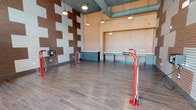 This single-floor bike room is designed to serve cyclists with both functionality and style. The space immediately draws attention with its striking feature wall, composed of alternating brown wood and white ribbed panels arranged in a bold geometric pattern that adds visual depth and a contemporary character to the room. Durable wood-style plank flooring runs throughout, providing a clean and practical surface that complements the overall design aesthetic. The room is equipped with multiple red freestanding bike repair stands, allowing cyclists to perform maintenance and adjustments with ease. An air station mounted to the feature wall provides convenient tire inflation. Along one wall, a long built-in workbench with teal cabinetry and ample drawer storage offers dedicated space for tools and bike-related tasks. The ceiling features exposed ductwork finished in a muted green-gray tone, lending an industrial touch, while pendant lights provide focused illumination throughout the space. Large glass entry doors flood the room with natural light and offer views of the surrounding outdoor environment. A secure storage unit near the entrance adds additional utility. The combination of purposeful amenities, thoughtful design details, and an open, well-lit layout makes this a highly functional and visually engaging space for cycling enthusiasts.
