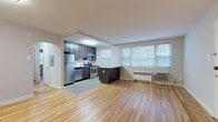 This single-floor residence is thoughtfully laid out across a well-proportioned floor plan, as illustrated by the overhead dollhouse view. Warm hardwood flooring flows consistently through the open living area and into both bedrooms, creating a cohesive and inviting atmosphere throughout the space.

The living area is bright and open, benefiting from a bank of windows along one wall that fill the room with natural light and offer outdoor views of surrounding trees. The kitchen is seamlessly integrated into the living space via a peninsula with dark granite countertops, and features dark espresso cabinetry, a subway tile backsplash, granite counters, and a full suite of stainless steel appliances, including a gas range, microwave, dishwasher, and refrigerator. A dedicated arched entryway connects the main living area to the rest of the unit.

Both bedrooms are comfortably sized, finished with the same warm hardwood flooring, neutral gray walls, ceiling fans, and windows that bring in natural light. Each room also includes baseboard heating units. The full bathroom is finished in classic white tile, with white tilework extending up the walls, a white vanity, and a window providing natural ventilation and light.
