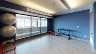 This single-level fitness studio is thoughtfully designed to support a wide range of training modalities. The space is defined by striking deep blue walls that create an energizing atmosphere, complemented by light wood-style flooring that adds warmth and durability underfoot. Large floor-to-ceiling windows line one side of the room, flooding the interior with natural light and offering views of the surrounding urban environment.

A generously sized wall mirror anchors one end of the studio, expanding the sense of space and allowing users to monitor their form during workouts. A wall-mounted flat-screen television is positioned above the mirror for entertainment or guided training sessions. Equipment is well-organized throughout: a double-tier kettlebell rack holds a full range of colorful weights, while stability balls are mounted neatly on the wall. A TRX suspension trainer is affixed to the wall alongside a rolled exercise mat, and red plyo boxes provide versatile options for strength and agility training.

The studio is accessed through a set of modern double glass-panel doors with a sleek frame, contributing to the open and connected feel of the space. Overhead linear lighting provides bright, even illumination throughout. This studio is a comprehensive and inviting space designed to accommodate functional fitness, strength training, and flexibility work.