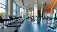 This contemporary, two-floor fitness center is defined by its impressive architectural design and extensive range of workout spaces. Towering floor-to-ceiling glass curtain walls flood the main cardio floor with abundant natural light while offering wide panoramic views of the surrounding outdoor landscape, including tree-lined streetscapes and neighboring buildings.

The primary cardio area accommodates a long row of treadmills, elliptical trainers, stationary bikes, and rowing machines, all positioned to take full advantage of the exterior views. Durable, speckled blue rubber flooring runs throughout, providing both a clean aesthetic and a functional, high-performance surface.

A dedicated strength and free weights zone features a well-curated selection of dumbbells, benches, and cable machines. This area is distinguished by warm wood slat accent walls backlit with ambient lighting, creating a bold visual contrast to the otherwise clean, minimalist palette. Full-length mirrors amplify the sense of space and assist with form during training.

The upper floor offers additional workout areas, including space for cycling equipment and functional training. Glass railings overlook the lower level, maintaining visual connectivity between floors. Linear LED lighting provides consistent, modern illumination throughout both levels, complementing the overall sleek and professional atmosphere of the facility.