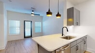 This thoughtfully designed one-bedroom, one-bathroom unit offers a well-organized single-floor layout that maximizes functionality and style. Light wood flooring flows consistently throughout every room, creating a warm and cohesive aesthetic.

The open-concept kitchen serves as the centerpiece of the living area, anchored by a spacious white quartz peninsula with an undermount sink and matte black faucet. Gray shaker-style cabinetry, a classic white tile backsplash, and a full suite of stainless steel appliances — including a refrigerator, dishwasher, microwave, and range — complete the modern kitchen. A pair of striking black pendant lights hang above the island, adding a contemporary focal point. A ceiling fan with integrated lighting serves the adjacent living area, which benefits from natural light through windows fitted with horizontal blinds.

The bedroom is a generously proportioned retreat featuring a ceiling fan, a wide window with natural light, and direct access to both the bathroom and a dedicated closet. The clean white walls and light flooring give the room an airy, calm atmosphere.

The bathroom is well-appointed with a gray vanity, a full soaking tub, and elegant marble-style tile surround with matte black fixtures, lending a refined, upscale feel to the space.