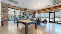 This single-floor amenity space is defined by its consistent use of reclaimed wood, which clads both the walls and ceiling throughout, creating a warm, rustic-industrial aesthetic. Polished concrete floors run seamlessly across the entire space, providing a sleek contrast to the textured wood surfaces. The layout is thoughtfully zoned into several distinct areas. A lounge area features tan leather armchairs arranged around a glass coffee table atop a bold patterned area rug, with a wall-mounted flat-screen TV flanked by vertical LED light fixtures. Adjacent to the lounge, a game area centers on a full-size pool table beneath woven pendant lights. Private dining booths with green upholstered seating line one wall, each equipped with individual TVs, offering semi-secluded spots for work or casual dining. A dedicated dining zone features a mid-century modern wood table beneath a striking starburst chandelier, positioned beside floor-to-ceiling glass walls that frame outdoor views of a pool area and surrounding buildings. A decorative patterned tile accent wall adds visual interest in this section. Large steel-framed windows and glass doors flood the interior with natural light throughout, reinforcing an open, connected atmosphere across the entire space.