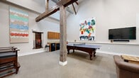 Located at 312 N 33rd St in Richmond, Virginia, this open-plan community game and recreation room showcases a striking blend of industrial heritage and modern amenity. The space is immediately defined by its dramatic vaulted ceilings, which soar high above the floor, supported by prominent exposed timber posts and crossbeams that lend a rustic, warehouse-inspired character to the bright, white-walled interior.

The generous, open floor plan is carpeted throughout and provides ample space to accommodate multiple activity zones. At the center of the room, a regulation-sized ping-pong table and an ornate dark-wood pool table serve as the primary focal points for recreation. A lounge area offers tufted leather armchairs arranged around a reclaimed-wood pallet coffee table on industrial caster wheels, creating a comfortable gathering spot. A wall-mounted flat-screen television and a vending machine add further convenience to this area.

Along one wall, a kitchenette includes upper wood cabinetry, a countertop, and under-counter refrigeration units, making the space suitable for social gatherings. Colorful large-scale abstract artwork adorns the walls, contributing an artistic energy to the room. Additional seating, including bar-height chairs, is available near a high-top table. The overall ambiance is welcoming, vibrant, and well-suited for community engagement and recreational use.
