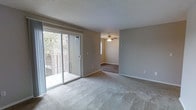 This single-floor space is designed with a straightforward, functional layout that connects a comfortable living area to an adjoining room. The main living space is finished with plush, neutral-toned carpeting that adds warmth underfoot, while the textured ceiling contributes a classic residential character. A prominent sliding glass door floods the area with natural light and provides direct access to a covered balcony, where views of surrounding trees create a pleasant outdoor connection.

The transition into the adjacent room is marked by a shift to darker hardwood-style flooring, offering a distinct visual contrast to the carpeted living area. This room is equipped with a ceiling fan, enhancing air circulation and adding a functional overhead fixture. A small window in this space supplements the natural light already introduced through the main living area's glass door.

Throughout the space, neutral beige walls provide a versatile backdrop suited to a variety of furnishing styles. Clean white baseboards frame each room, contributing to a tidy, well-maintained appearance. The overall ambiance is calm and understated, making the space an adaptable and practical environment.