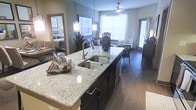 This single-floor residence showcases a thoughtfully designed open-concept layout that connects the kitchen, dining, and living areas into one cohesive space. The kitchen serves as the centerpiece, anchored by a large granite-topped island with a double stainless steel undermount sink and elegant pendant lighting hanging overhead. Dark espresso cabinetry provides a striking contrast against the light granite countertops, while rich, dark hardwood flooring extends throughout the space, adding warmth and continuity.

Natural light fills the interior through multiple windows dressed with white blinds, creating a bright and welcoming atmosphere. The dining area is positioned near a glazed exterior door, offering an easy connection to the outdoors. Beyond the kitchen, the living area flows naturally, providing ample room for comfortable furnishings.

A mirrored entryway near the kitchen reflects the adjacent bedroom, which features coordinated décor and soft, neutral tones. The overall ambiance is modern and refined, with carefully chosen finishes and fixtures that balance style and function throughout the space.