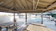 This outdoor community amenity space is centered around a generously sized swimming pool enclosed by white fencing, offering a clean and inviting aquatic retreat. A prominent covered pavilion anchors the area, featuring an exposed wood beam and rafter ceiling supported by sturdy columns. The vaulted structure provides ample shade over a concrete patio, making it a comfortable gathering spot regardless of the weather. Beneath the pavilion, multiple metal picnic tables with attached bench seating offer space for dining and socializing. Adjacent to the seating area, a designated BBQ zone is equipped with charcoal grills mounted on masonry platforms, along with a metal waste receptacle — ideal for outdoor cooking and entertaining. The perimeter of the space is bordered by a landscaped hillside with low-growing shrubs and mature trees, adding a natural backdrop to the recreational setting. Yellow safety markings accent the concrete pathways, clearly delineating accessible routes throughout the area. The overall layout is thoughtfully designed for communal use, combining functional amenities with an open, relaxed atmosphere well-suited for casual outdoor living.