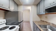 This single-floor unit offers a functional layout across two bedrooms, one bathroom, a hallway, and a kitchen. The layout is efficiently organized, with the galley-style kitchen positioned near the entry. The kitchen features gray cabinetry, laminate countertops, a stainless refrigerator, a white electric range, and a built-in sink — all arranged along a narrow corridor that leads into the main living areas. An intercom panel is mounted near the kitchen entry wall, adding a practical convenience feature.

The primary bedroom is the most spacious room in the unit, offering generous floor area covered in neutral carpeting. It features a large window dressed with vertical blinds, a wall-mounted air conditioning unit, and baseboard heating — providing year-round climate control. The second bedroom is comparably sized, also carpeted, with its own window and baseboard heating.

The bathroom features full wall-to-ceiling dark brown ceramic tile with accent detailing, a white soaking tub with shower, a wall-mounted sink, and mosaic tile flooring — giving it a distinctly retro aesthetic. Throughout the unit, neutral tones, carpet flooring, and panel ceilings contribute to a straightforward, functional ambiance.