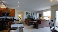 This single-floor space showcases a thoughtfully designed open-concept layout that integrates the kitchen and living area into one cohesive, functional environment. The kitchen features warm wood cabinetry, a sleek dark countertop with a built-in sink, and bar-height seating, creating an ideal casual dining and entertaining space. Recessed lighting throughout the kitchen and living area ensures consistent, even illumination across the entire space.

The living room is generously proportioned, with plush wall-to-wall carpeting underfoot and neutral-toned walls that lend a calm, welcoming ambiance. Multiple windows, dressed with adjustable blinds, allow for an abundance of natural light while maintaining privacy. The room comfortably accommodates two large sofas arranged around a central media console, which houses a flat-screen television flanked by a curated gallery of framed artwork.

Decorative touches such as accent lamps, a lantern-style floor piece, and a cozy throw blanket add warmth and personality to the living space. A hallway visible from the main area suggests additional rooms beyond, rounding out the functional floor plan. Overall, the space offers a comfortable, modern aesthetic with a practical and inviting layout.