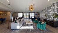 This single-floor space is designed as a stylish communal gathering area that seamlessly blends functionality with contemporary aesthetics. The open-plan layout flows naturally from a lounging zone into a dining area and a well-appointed kitchenette, creating a versatile environment suited for both relaxed socializing and organized events.

The lounge area is a focal point of the space, featuring a plush gray sofa, bold emerald-green velvet accent chairs, and a marble-topped coffee table anchored on a textured area rug. A striking gold sputnik chandelier overhead adds a dramatic, upscale touch. Large windows dressed with two-toned navy and white floor-length drapes flood the interior with abundant natural light, while offering views of an outdoor patio furnished with seating.

The dining zone accommodates a long table surrounded by modern blue upholstered chairs, providing a defined area for group gatherings. Adjacent to this is the kitchenette, which features rich walnut cabinetry, a white quartz countertop, a stainless steel sink, and a black microwave. A bold geometric tile accent wall adds visual interest and texture behind the counter.

Throughout, light wood-look flooring unifies the space, while gold-framed mirrors and curated décor contribute to an overall atmosphere that is polished, welcoming, and contemporary.
