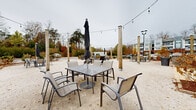Located at 485 Crafton Cir in Charlottesville, Virginia, this outdoor communal space is designed for social gatherings and relaxed outdoor living. The ground-level area features a compacted gravel surface that provides a casual, open-air atmosphere throughout. Wooden posts are anchored at regular intervals across the space, supporting overhead string lights that create an inviting ambiance during evening hours. Multiple seating arrangements are distributed throughout the area, including square patio tables paired with sling-style chairs and umbrella stands, offering comfortable spots for dining or conversation. A central fire pit surrounded by additional seating serves as a natural focal point for the space. A built-in outdoor kitchen station equipped with multiple grills provides a convenient amenity for outdoor cooking and entertaining. Landscaped greenery and mature trees frame the perimeter, offering a natural backdrop, while views of the surrounding residential community are visible from all sides. Pole-mounted lights supplement the string lighting for added visibility. The open layout accommodates a variety of uses, from casual gatherings to larger community events, making it a versatile and well-appointed outdoor amenity.
