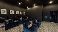 This single-floor screening room is thoughtfully designed to deliver a premium cinematic experience within a refined, lounge-style setting. The space is enveloped in deep charcoal and matte black finishes across the walls and ceiling, creating an immersive, theater-like atmosphere that minimizes light interference and enhances viewing conditions.

A ceiling-mounted projector delivers content onto a large, centrally positioned projection screen framed by built-in shelving units on either side, which are styled with decorative objects and books. Recessed lighting provides subtle, controlled illumination throughout the room. The main seating area features generously sized, deep-blue velvet lounge chairs and sofas arranged in rows across a textured area rug, each accompanied by small side tables for added convenience.

Toward the rear of the room, a raised bar area offers high-top tables paired with upholstered barrel chairs, creating a casual viewing perch beneath a curated gallery of four framed abstract artworks. A large sliding glass door along one wall allows natural light to filter in when desired, with roller shades available for full light control. The overall ambiance is intimate, upscale, and purpose-built for group entertainment.