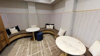 This thoughtfully designed lounge space exudes a sophisticated yet comfortable atmosphere through a careful blend of textures and finishes. At its heart is a generously curved banquette, upholstered in warm taupe and framed by striking gold-trimmed paneling that lines the lower half of the walls. Above the trim, the walls feature vertical shiplap-style paneling in a soft white, lending a refined, contemporary character to the room.

Two circular marble-topped tables with brushed gold pedestal bases anchor the seating area, providing functional surfaces while reinforcing the space's upscale aesthetic. An assorted mix of cushions in cream, white, and navy adds layered comfort to the banquette. A navy cylindrical ottoman introduces a bold accent that complements the overall neutral palette.

The flooring is covered by a plush area rug in warm beige with a subtle geometric pattern, softening the space and defining the seating zone. Overhead, a recessed ceiling detail adds architectural interest and a sense of depth. The overall ambiance is intimate and polished, well-suited for socializing or quiet relaxation.