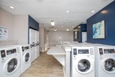 Located at 3800 Club Dr in Norcross, Georgia, this well-appointed community laundry facility offers a clean, modern environment designed for convenience and ease of use. The single-floor space features a contemporary aesthetic with a neutral gray and bold navy blue color palette, wood-look flooring, and recessed lighting that keeps the room bright and welcoming throughout.

The facility is equipped with multiple front-loading washing machines and stacked dryer units, providing ample capacity to serve a large number of residents. Instructional washing and drying guides are posted on the walls for easy reference. Centrally positioned folding counters with light-toned surfaces run the length of the room, offering generous workspace for sorting and folding laundry.

Large windows along the perimeter allow natural light to fill the space and provide views of the surrounding greenery, enhancing the overall atmosphere. Decorative artwork on the navy accent walls adds a touch of character, elevating the space beyond a purely utilitarian function. The layout is thoughtfully organized to allow comfortable movement between machines and folding areas, making the laundry experience as efficient and pleasant as possible.