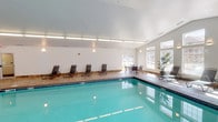 Located at 1445 W Gateway Cir S in Fargo, North Dakota, this single-floor indoor pool facility offers a welcoming and well-appointed aquatic environment. The centerpiece of the space is a generously sized rectangular swimming pool with clear, blue-green water, surrounded by tile and deck flooring that provides ample room for movement and relaxation.

The pool area is designed with comfort in mind, featuring multiple lounge chairs positioned along the poolside for relaxing before or after a swim. A seating area with a table and chairs is also present, offering a casual spot to gather or unwind. Decorative touches, including a potted tropical plant, add a refreshing, resort-like feel to the interior.

The room is bright and airy, benefiting from an abundance of natural light that streams in through large, multi-pane windows lining the walls. The vaulted ceiling with recessed linear lighting enhances the sense of openness and height, contributing to the facility's clean and modern aesthetic. The white walls and light-toned surfaces throughout maintain a crisp, inviting atmosphere.

This indoor pool space is well-suited for year-round use, a particularly valuable amenity given Fargo's climate, providing a comfortable and functional environment for swimming and leisure activities regardless of the season.
