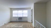This single-floor space offers a clean, functional layout with a straightforward and practical design. The main living area is characterized by neutral beige walls and light-toned carpeted flooring, creating a simple and welcoming atmosphere. A wide, multi-pane window dominates one wall, allowing generous natural light to fill the room while offering views of the surrounding exterior. Climate comfort is addressed through a wall-mounted air conditioning unit paired with baseboard heating, ensuring year-round temperature control. A classic six-panel white interior door provides access to an adjoining room, maintaining a cohesive and traditional aesthetic throughout. A notable architectural detail is the decorative white metal staircase railing visible along one edge of the space, adding a subtle ornamental touch to the otherwise understated interior. The open floor plan lends itself to flexible furniture arrangements, making the space adaptable to a variety of uses. Overall, the space presents a practical and well-lit environment with essential amenities and a clean, no-frills design sensibility.