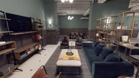 This thoughtfully designed single-floor space blends industrial character with refined, eclectic style. The interior is anchored by deep sage green walls accented with classic dark wood wainscoting, creating a warm and sophisticated atmosphere throughout. Exposed ceiling beams and visible ductwork reinforce the industrial aesthetic, while a combination of pendant and wall-mounted lighting adds warmth and dimension to the space.

The layout is divided into distinct functional zones that flow seamlessly into one another. A comfortable lounge area features a plush teal velvet sofa paired with an upholstered ottoman and a sleek coffee table, all set atop a patterned area rug over classic mosaic tile flooring. Leather accent chairs and layered textiles add depth and a curated, lived-in feel.

Toward the rear, a dedicated dining or meeting area accommodates a full-sized table surrounded by leather chairs, ideal for group gatherings or collaborative work sessions. Open brass-framed shelving units along the perimeter display books, décor, and curated accessories, reinforcing the space's library-like sensibility.

Overall, the space strikes a balance between functionality and design-forward aesthetics, making it well-suited as a communal lounge, co-working environment, or private event venue.