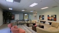 This single-floor space is thoughtfully designed to serve as both a recreational hub and a relaxed lounge area. The open layout accommodates a full-size ping pong table alongside a comfortable seating arrangement, making it well-suited for both active play and casual gatherings. The lounge area features upholstered chairs and low-profile coffee tables arranged atop a striped area rug, creating a welcoming focal point within the room. Decorative vases and wall-mounted artwork add visual interest and a sense of curated style throughout the space. The walls are painted in a soft teal-gray tone that lends a calm, contemporary ambiance, complemented by crisp white trim and baseboards. A large mirror on one wall enhances the sense of openness, reflecting light and visually expanding the room. The drop ceiling is fitted with recessed fluorescent panel lighting that provides even, bright illumination throughout. Neutral-toned carpet covers the main floor area, contributing to the overall comfortable and inviting atmosphere. A door at the rear of the space provides additional access, and subtle signage suggests this is a shared or amenity-style environment designed for community use and relaxation.