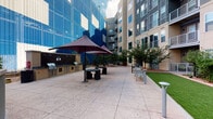 This expansive outdoor courtyard is a thoughtfully designed amenity space nestled within the interior of a contemporary multi-story residential complex. The space is defined by wide, light-colored concrete pavers that create a clean, open atmosphere throughout. A dedicated outdoor kitchen and BBQ station, built with brick surround and equipped with multiple grills, provides an ideal setting for gatherings and al fresco dining. Large market-style umbrellas shade circular bistro tables and stools nearby, offering a comfortable place to dine outdoors. Scattered throughout the courtyard are modern metal benches and high-top seating areas, encouraging casual interaction. A standout feature is the sunken fire pit lounge, bordered by a curved low wall and surrounded by contemporary lounge chairs — a welcoming focal point for relaxed evenings. In a separate zone, vibrant teal chaise lounge chairs are arranged near striking blue architectural cladding, adding a bold pop of color. Manicured turf lawn areas, mature trees, ornamental plantings, and landscaped garden beds soften the hardscape and introduce natural texture. Pathway lighting throughout ensures the space is both functional and inviting after dark. The surrounding building facades, complete with balconies and large windows, frame the courtyard and contribute to its sheltered, community-oriented character.
