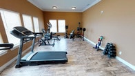 This dedicated fitness room offers a well-rounded workout environment designed to accommodate a variety of exercise routines. The space is finished with warm golden-tan walls and elegant crown molding, lending it a polished appearance that sets it apart from a typical gym setting. Wood-look tile flooring runs throughout, providing both durability and a clean aesthetic that complements the overall design.

The room is generously sized, offering ample floor space to move freely between equipment without feeling cramped. Cardio equipment includes a treadmill and an elliptical trainer, positioned to take advantage of natural light streaming through multiple windows dressed with white blinds. An upright stationary bike adds further cardio variety.

For strength training, the room features a weight bench with a barbell rack, a selection of medicine balls, and a dedicated kettlebell storage stand holding multiple weights of varying sizes. Additional free weights, including dumbbells, are neatly organized along the wall. Flush-mount ceiling lights provide consistent overhead illumination throughout the space.

Overall, the fitness room strikes a balance between functional workout utility and refined interior finishes, making it a practical and inviting space for daily exercise.