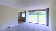 This single-story home offers a practical and comfortable layout designed for easy living. The main living area is finished with plush carpeting underfoot and neutral-toned walls that create a calm, understated atmosphere. A standout feature of the living space is the expansive sliding glass door wall, which floods the interior with natural light and provides unobstructed views of a well-maintained green lawn and surrounding trees — offering a seamless visual connection to the outdoors. The sliding doors also provide direct access to the exterior, making outdoor entertaining and everyday living convenient. Adjacent to the main living area, a separate room — likely a dining area — is visible through a wide entryway and is accentuated by a hanging pendant light fixture, adding a touch of character. A window in this secondary space allows additional natural light to filter through. A rustic shingle-clad accent wall near the sliding doors adds a distinctive architectural detail, lending texture and visual interest to the otherwise clean interior. The overall ambiance of the home is relaxed and functional, with an emphasis on natural light, open sightlines, and a strong connection to the surrounding landscape.