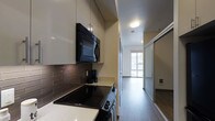 This single-floor space showcases a thoughtfully designed interior that balances functionality with contemporary style. The kitchen is outfitted with glossy white upper cabinetry, light-toned countertops, and a dark subway tile backsplash that adds visual contrast. Black appliances, including an over-the-range microwave and a smooth-top electric range, contribute to a cohesive, modern aesthetic. The kitchen layout is efficient, maximizing storage and workspace within a streamlined footprint. A flush ceiling light provides direct illumination to the cooking area. Adjacent to the kitchen, a hallway with mirrored sliding doors — likely concealing a closet — connects to the main living area. The living space benefits from natural light entering through a large window, creating a bright and open ambiance. Hardwood-style flooring runs consistently throughout, lending warmth and visual continuity to the unit. The overall design emphasizes clean lines, neutral tones, and practical use of space, making it a well-appointed and functional single-level residence.